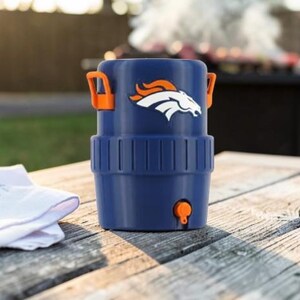 May include: A navy blue drinks cooler with orange handles and a tap. The cooler displays the Denver Broncos logo in white and orange. It rests on a weathered wooden table, with a white cloth nearby.