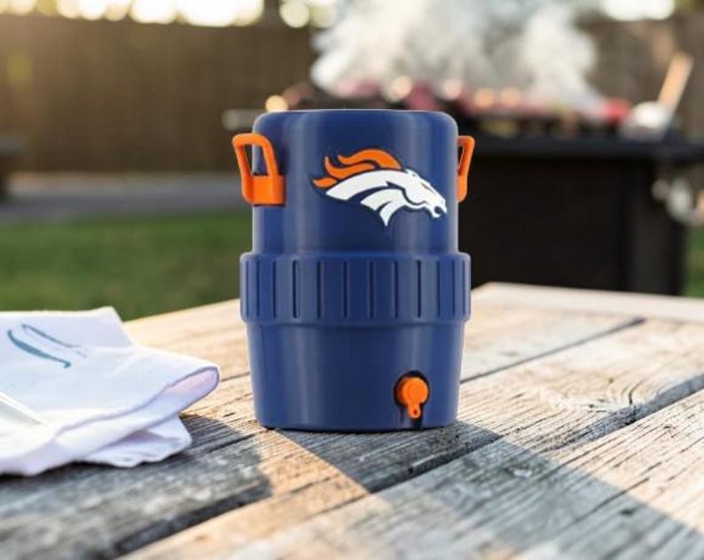 May include: A navy blue drinks cooler with orange handles and a tap. The cooler displays the Denver Broncos logo in white and orange. It rests on a weathered wooden table, with a white cloth nearby.