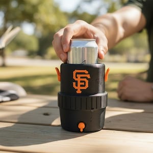 Pode incluir: Refrigerador de latas isolado preto e laranja com o logotipo do San Francisco Giants. O refrigerador tem al&ccedil;as e uma torneira laranja. Uma lata prateada &eacute; colocada dentro. Em uma mesa de piquenique de madeira.