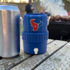 May include: A silver drinks can stands beside a blue drinks cooler featuring the Houston Texans logo. The cooler has a white tap and handles. The items are on a weathered wooden picnic table, with a barbecue and people in the background.