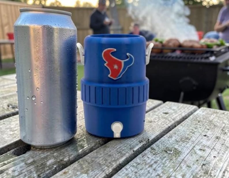 May include: A silver drinks can stands beside a blue drinks cooler featuring the Houston Texans logo. The cooler has a white tap and handles. The items are on a weathered wooden picnic table, with a barbecue and people in the background.