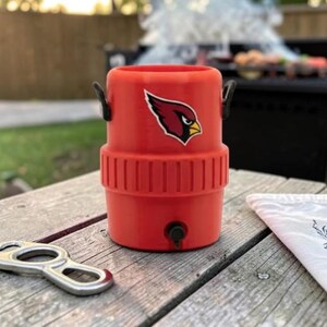 May include: A red drinks cooler with a black handle and a team logo. A silver bottle opener lies on a wooden table next to a white cloth. A barbecue grill is in the background.