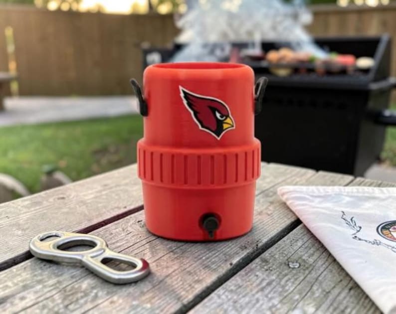 May include: A red drinks cooler with a black handle and a team logo. A silver bottle opener lies on a wooden table next to a white cloth. A barbecue grill is in the background.