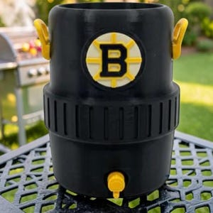May include: Black beverage cooler with yellow accents, featuring the Boston Bruins logo. The cooler has a spigot and two handles. It is sitting on a metal table outdoors, with a grill in the background.