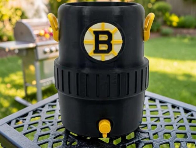 May include: Black beverage cooler with yellow accents, featuring the Boston Bruins logo. The cooler has a spigot and two handles. It is sitting on a metal table outdoors, with a grill in the background.