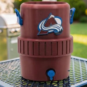 May include: A maroon water cooler with blue handles and a blue spigot. The cooler features the Colorado Avalanche logo in white, blue, and maroon. The cooler is sitting on a black metal table.