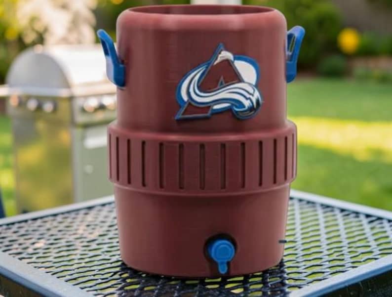 May include: A maroon water cooler with blue handles and a blue spigot. The cooler features the Colorado Avalanche logo in white, blue, and maroon. The cooler is sitting on a black metal table.
