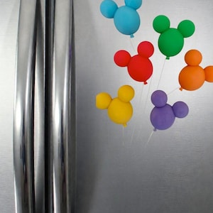 May include: A collection of colorful, Mickey Mouse-shaped balloons in blue, red, green, orange, yellow, and purple. The balloons are attached to thin strings and appear to be floating against a silver refrigerator door. The balloons are a fun, whimsical decoration.