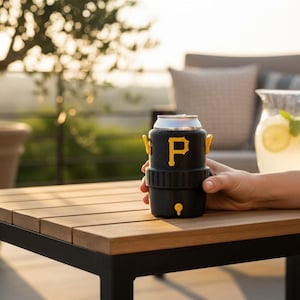 Pode incluir: Um refrigerador de latas preto com detalhes amarelos e a letra "P" amarela. O refrigerador est&aacute; segurando uma lata prateada. O refrigerador est&aacute; em uma mesa de madeira. Uma jarra de limonada est&aacute; ao fundo.
