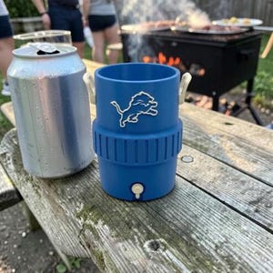May include: A blue insulated drinks holder with a white Detroit Lions logo, next to a silver can on a wooden picnic table. The holder has a spigot and white handles. A grill is in the background.