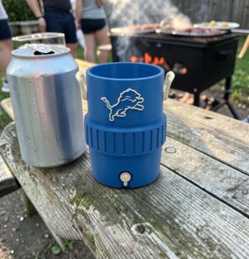May include: A blue insulated drinks holder with a white Detroit Lions logo, next to a silver can on a wooden picnic table. The holder has a spigot and white handles. A grill is in the background.
