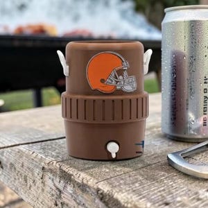 May include: A brown drinks cooler featuring a Cleveland Browns helmet logo, beside a silver can of soda and a bottle opener. The cooler has white handles and a small tap. The items are on a weathered wooden surface.