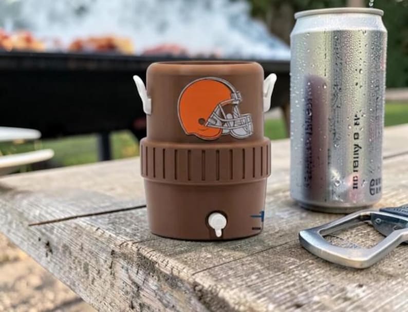 May include: A brown drinks cooler featuring a Cleveland Browns helmet logo, beside a silver can of soda and a bottle opener. The cooler has white handles and a small tap. The items are on a weathered wooden surface.
