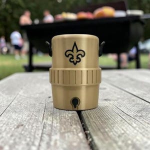 May include: A gold-coloured drinks container featuring a black fleur-de-lis logo. The container has a cylindrical shape with a handle on each side. It is on a wooden picnic table. The background shows a barbecue and people.