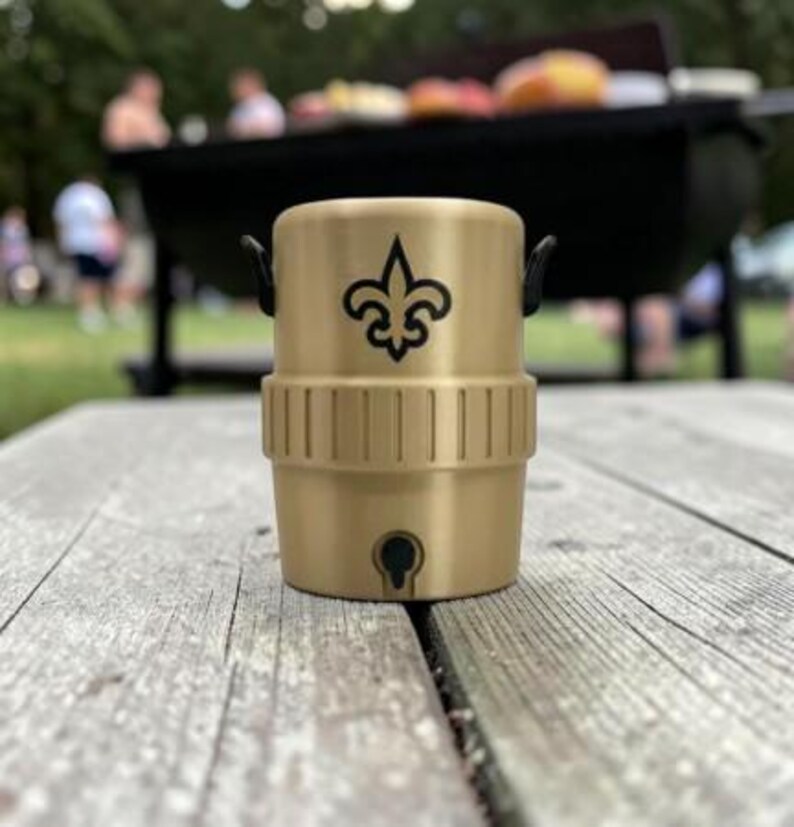 May include: A gold-coloured drinks container featuring a black fleur-de-lis logo. The container has a cylindrical shape with a handle on each side. It is on a wooden picnic table. The background shows a barbecue and people.