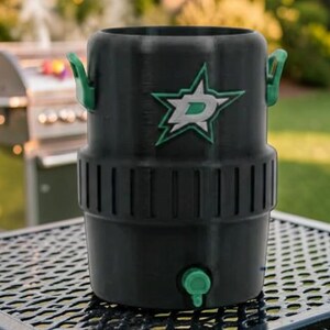 May include: Black insulated beverage cooler with green handles and a green spigot. The cooler features a green and white logo of a star with a letter "D" inside. The cooler is sitting on a metal table.