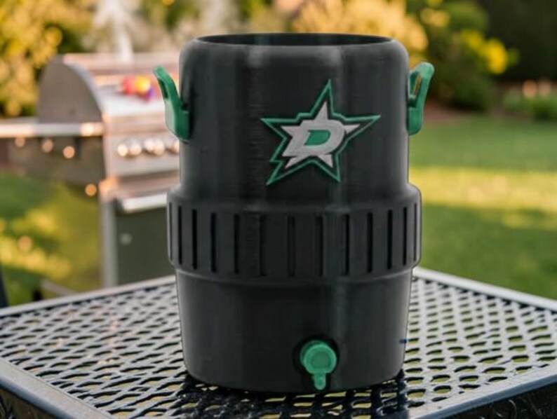 May include: Black insulated beverage cooler with green handles and a green spigot. The cooler features a green and white logo of a star with a letter "D" inside. The cooler is sitting on a metal table.