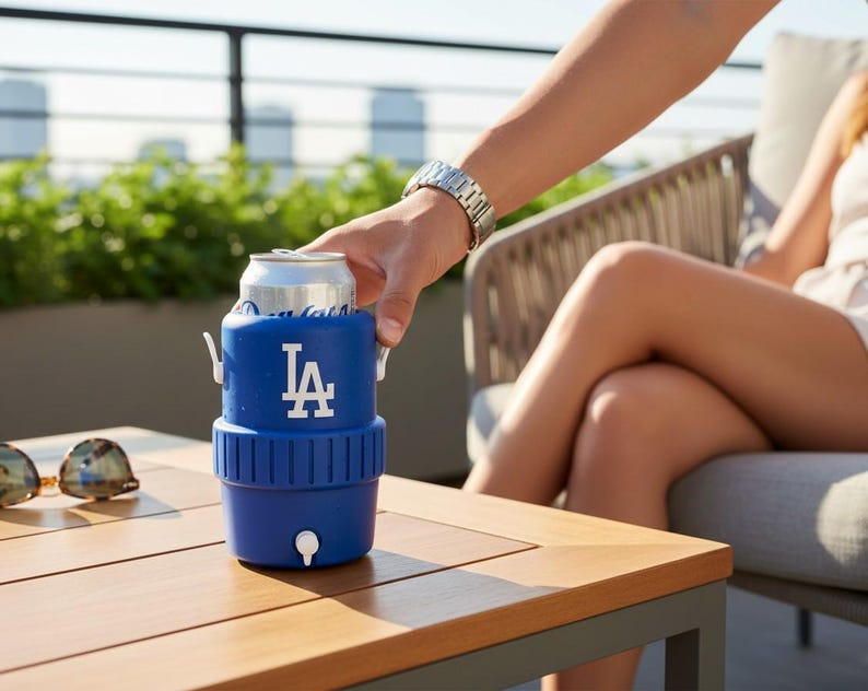 Pode incluir: Um refrigerador de latas azul com o logotipo dos Dodgers de Los Angeles em branco. O refrigerador est&aacute; segurando uma lata prateada e tem uma pequena torneira branca. O refrigerador est&aacute; em uma mesa de madeira.