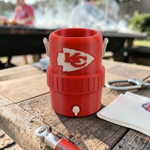 May include: A red insulated drinks holder featuring a white logo of a team's arrowhead and the letters "KC". The holder has a small white tap and two white handles. It sits on a weathered wooden table.