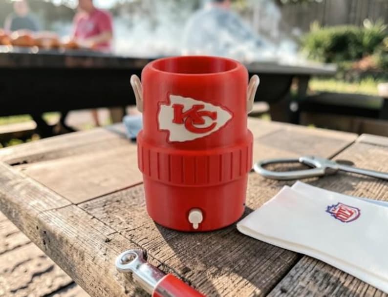 May include: A red insulated drinks holder featuring a white logo of a team's arrowhead and the letters "KC". The holder has a small white tap and two white handles. It sits on a weathered wooden table.