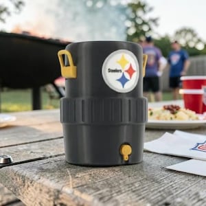May include: A black insulated drinks cooler with yellow handles, displaying the Pittsburgh Steelers logo. The cooler is on a wooden picnic table, with a barbecue and people in the background. A red cup and food are also visible.