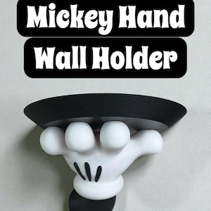 May include: A white and black Mickey Mouse hand wall holder. The holder features a black top and a white hand with black details. The text "Mickey Hand Wall Holder" is displayed in white on a black background above the holder.