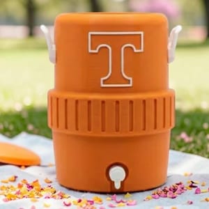 May include: Orange water cooler with a white "T" logo. The cooler has a spigot, white handles, and a matching lid. It sits on a blue and white patterned cloth, with flower petals scattered around it.