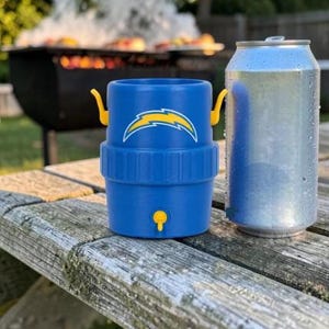 May include: A blue drinks cooler with a yellow and white logo, next to a silver can. The cooler has yellow handles and a yellow tap. The background shows a barbecue grill with food and smoke, suggesting an outdoor event.