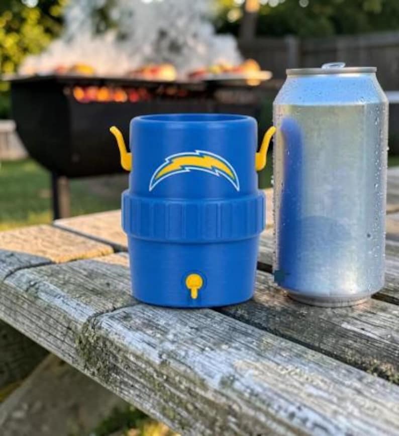 May include: A blue drinks cooler with a yellow and white logo, next to a silver can. The cooler has yellow handles and a yellow tap. The background shows a barbecue grill with food and smoke, suggesting an outdoor event.