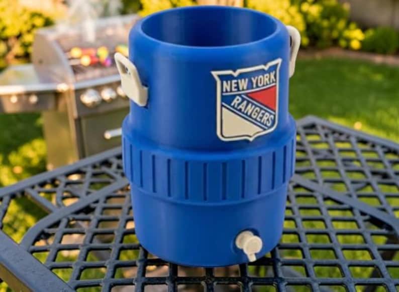 May include: A blue beverage cooler with the New York Rangers logo. The cooler has a spigot, white handles, and a ribbed design. It is sitting on a black metal table outdoors.