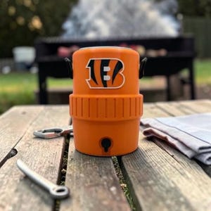 May include: An orange cooler featuring the Cincinnati Bengals logo, a black handle, and a tap. A bottle opener and a tea towel rest on the wooden table. A barbecue grill is in the background.