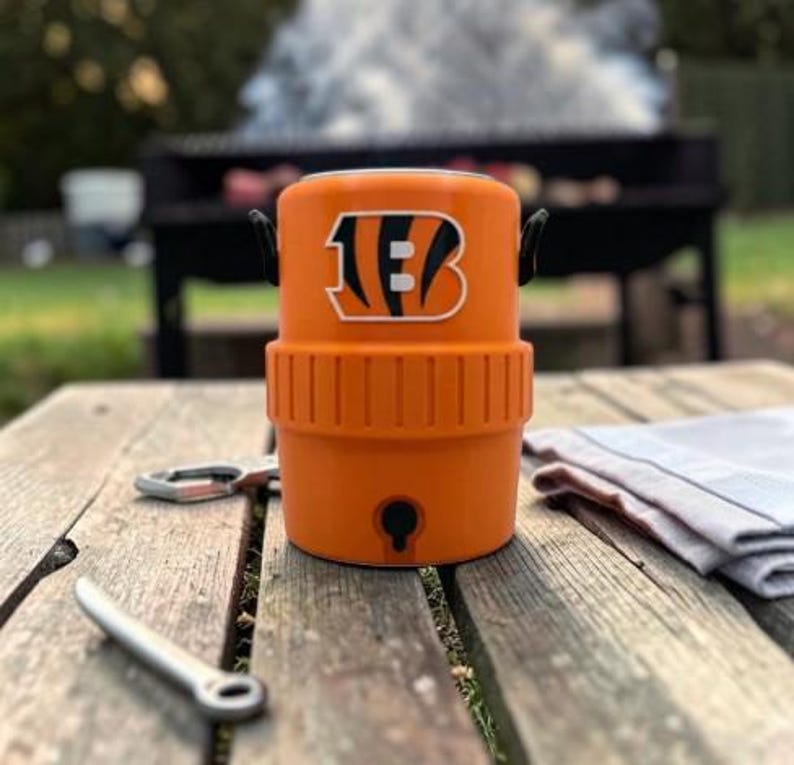 May include: An orange cooler featuring the Cincinnati Bengals logo, a black handle, and a tap. A bottle opener and a tea towel rest on the wooden table. A barbecue grill is in the background.