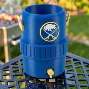May include: A blue beverage cooler with a yellow spigot and handles. The cooler features a gold and white emblem with a buffalo and crossed swords. The cooler is sitting on a black metal table with a grill in the background.