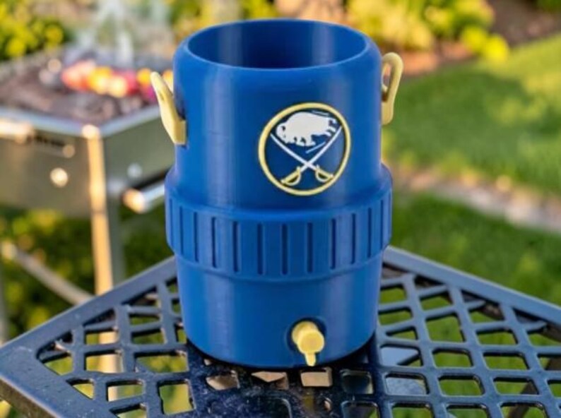 May include: A blue beverage cooler with a yellow spigot and handles. The cooler features a gold and white emblem with a buffalo and crossed swords. The cooler is sitting on a black metal table with a grill in the background.