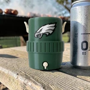 May include: A green drinks cooler featuring the Philadelphia Eagles logo, beside a silver can with condensation. The cooler has white handles and a small white tap. The items are on a weathered wooden picnic table.