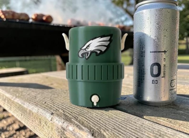 May include: A green drinks cooler featuring the Philadelphia Eagles logo, beside a silver can with condensation. The cooler has white handles and a small white tap. The items are on a weathered wooden picnic table.