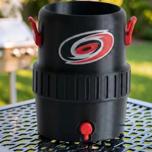 May include: Black beverage cooler with red accents, featuring a team logo. The cooler has a cylindrical shape with a red spigot and handles. It is sitting on a metal table outdoors.