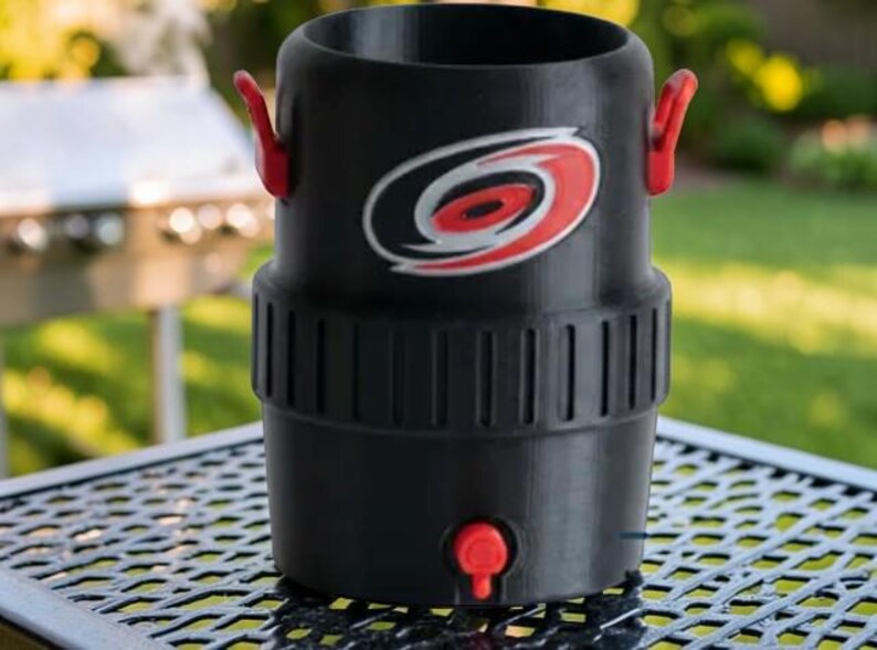 May include: Black beverage cooler with red accents, featuring a team logo. The cooler has a cylindrical shape with a red spigot and handles. It is sitting on a metal table outdoors.