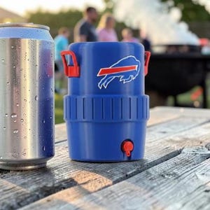 May include: A silver drinks can with condensation next to a blue drinks cooler with red handles and a team logo. The cooler has a red tap and is on a weathered wooden picnic table.