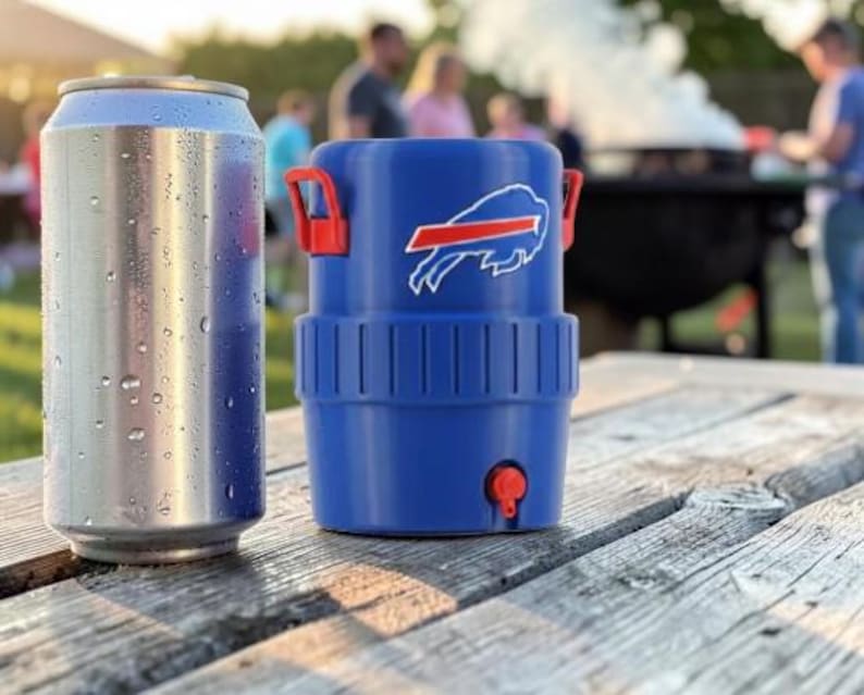 May include: A silver drinks can with condensation next to a blue drinks cooler with red handles and a team logo. The cooler has a red tap and is on a weathered wooden picnic table.