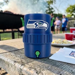 May include: A blue insulated drinks cooler featuring the Seattle Seahawks logo. The cooler has green handles and a tap. It's on a wooden picnic table with food and drinks, suggesting an outdoor event.