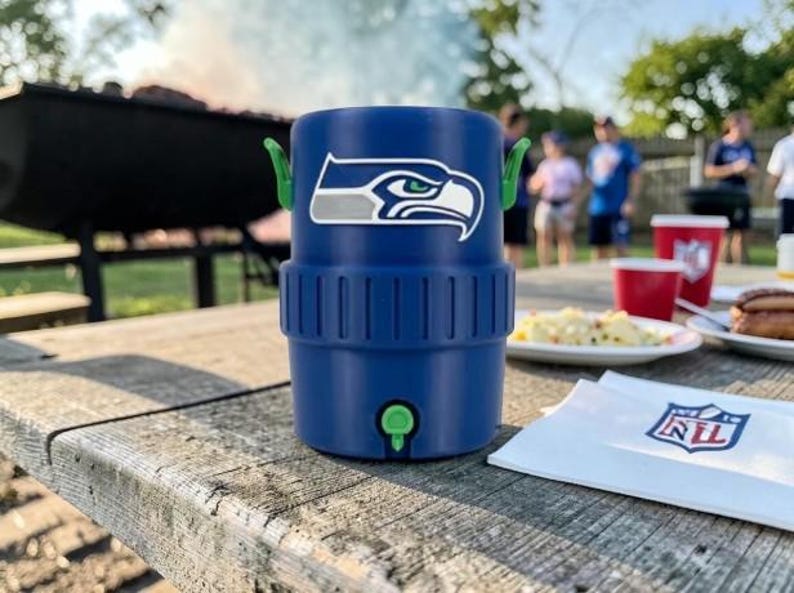 May include: A blue insulated drinks cooler featuring the Seattle Seahawks logo. The cooler has green handles and a tap. It's on a wooden picnic table with food and drinks, suggesting an outdoor event.