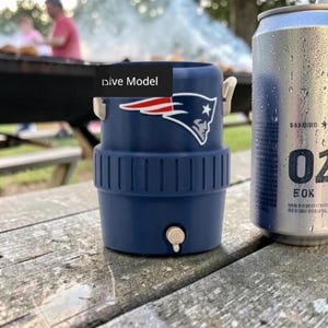 May include: A navy blue drinks cooler with the New England Patriots logo next to a silver can with condensation. The cooler has a white tap and handles. The can has the number "02" printed on it.