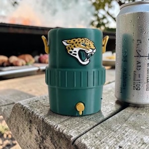 May include: A green drinks cooler featuring the Jacksonville Jaguars logo, beside a can of soda. The cooler has a yellow handle and a small yellow tap. The can is silver with condensation.