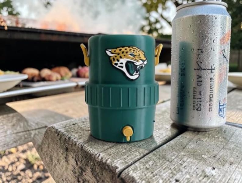 May include: A green drinks cooler featuring the Jacksonville Jaguars logo, beside a can of soda. The cooler has a yellow handle and a small yellow tap. The can is silver with condensation.