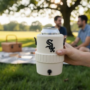 Pode incluir: Um refrigerador de latas creme com um logotipo preto, segurando uma lata prateada. O refrigerador tem al&ccedil;as pretas e uma pequena torneira na parte inferior. A imagem &eacute; ao ar livre com uma cesta de piquenique e pessoas ao fundo.
