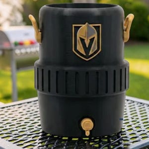 May include: Black beverage cooler with gold-colored accents, including handles and a spigot. The cooler features the Vegas Golden Knights logo in gold. It is sitting on a metal table outdoors, with a grill in the background.