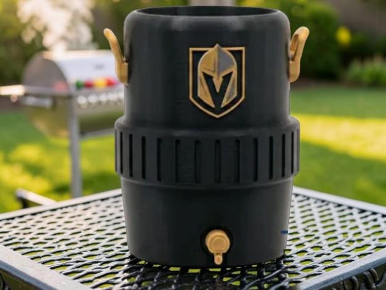 May include: Black beverage cooler with gold-colored accents, including handles and a spigot. The cooler features the Vegas Golden Knights logo in gold. It is sitting on a metal table outdoors, with a grill in the background.