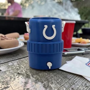 May include: A blue insulated drinks holder with a white horseshoe logo, a white tap, and white handles. The holder is on a wooden picnic table with food and a red cup in the background. A napkin with an NFL logo is also on the table.