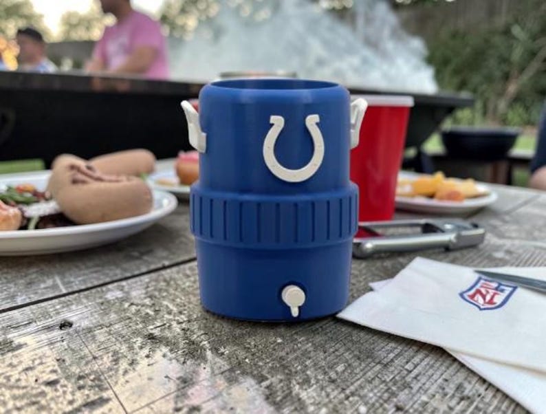 May include: A blue insulated drinks holder with a white horseshoe logo, a white tap, and white handles. The holder is on a wooden picnic table with food and a red cup in the background. A napkin with an NFL logo is also on the table.